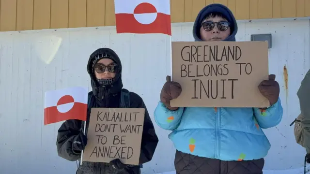 Двое в теплых куртках, солнечных очках и рукавицах стоят с гренландскими флажками и написанными на картонках словами "Гренландия принадлежит инуитам", "Гренландия не хочет быть аннексированной"