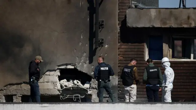 Quatro homens com coletes da polícia, bem como um homem com uma roupa branca contra materiais perigosos, capacete e máscara facial, são vistos ao lado de um prédio que tem um buraco na lateral após o ataque do drone