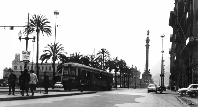 Foto em preto e branco de Barcelona em 1969, com uma rua, um ônibus passando, várias pessoas atravessando e, ao fundo, a estátua de Colombo.