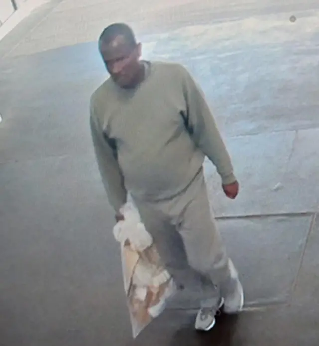 灰色のトレーナーとトレパンにスニーカーと言う服装で、右手に茶色い紙などが入った透明のプラスチックバッグを手に歩道を歩くアフリカ系の男性