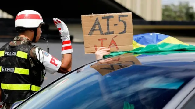 Guarda ao lado de carro com pessoa segurando placa que diz 'AI-5 já'