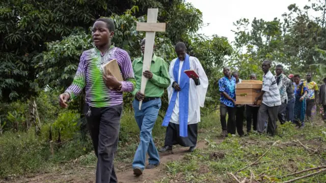 Un pastor camina al frente de quienes llevan los féretros de madera y un cruz de madera