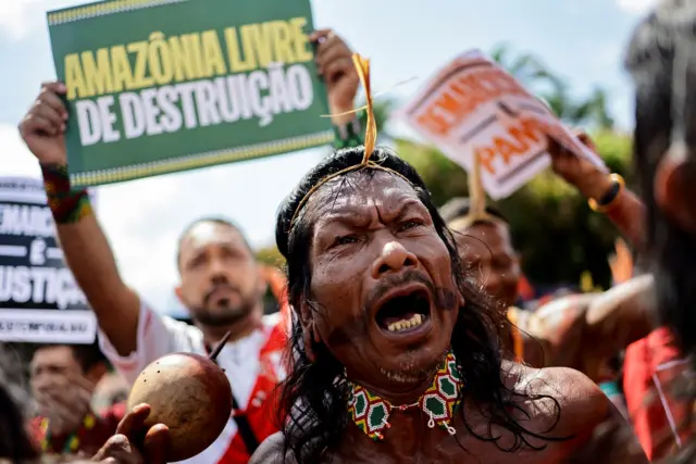 Indígena gritandodownload esporte betprotesto, com cartazes atrás pedindo proteção à Amazônia