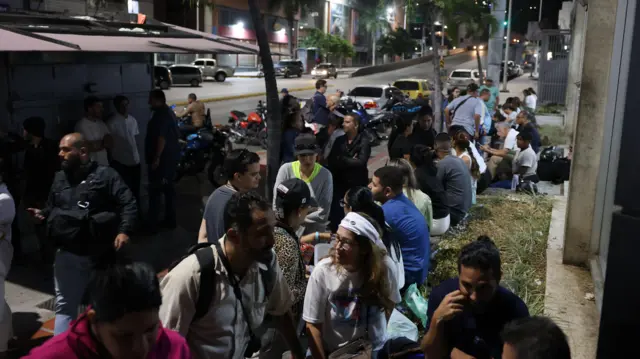 Темная улица, возле одного из зданий собралось множество людей, некоторые сидят на бордюре у газона возле обочины. Вдоль дороги посажены пальмы и припаркованы машины.