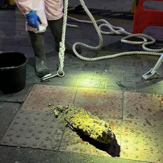 Pessoa com roupas de proteção, segurando uma corda, aponta para um pedaço de gordura solidificada retirado do esgoto