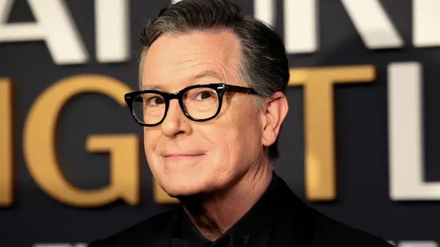 Stephen Colbert, presentador de Late Show, mira a la cámara con una leve sonrisa. Tiene gafas de montura negra.