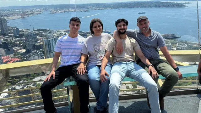Una familia de cuatro se encuentra en un edificio alto con ventanas que dan a la ciudad y al mar. Hay dos jóvenes: los hijos, la madre y el padre. Todos tienen el pelo oscuro y están sentados juntos, sonriendo.