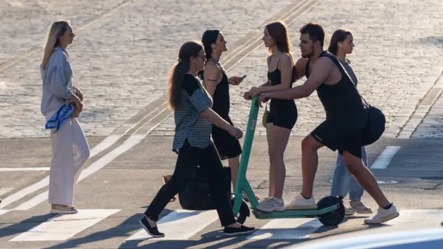 Хлопець із дівчиною вдвох переїжджають проїжджу частину по зебрі на самокаті, навколо них іде багато людей