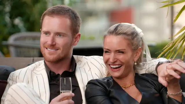 Dos participantes del programa de telerrealidad de Netflix sentados en un sofá y agarrados de las manos mientras sonríen y miran a un costado de la cámara