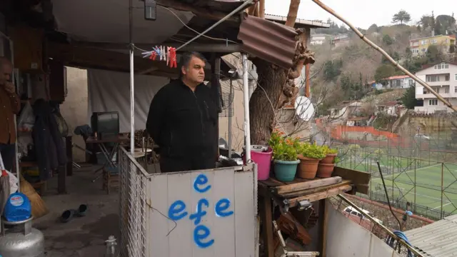 Fotoğrafta Adem Demir, Sarıyer'deki evinin bahçesinde görülüyor. Demir, uzağa doğru bakıyor. Ayakta durduğu yerin önündeki plakada Efe yazıyor. Arkada uzakta evler gözüküyor. 