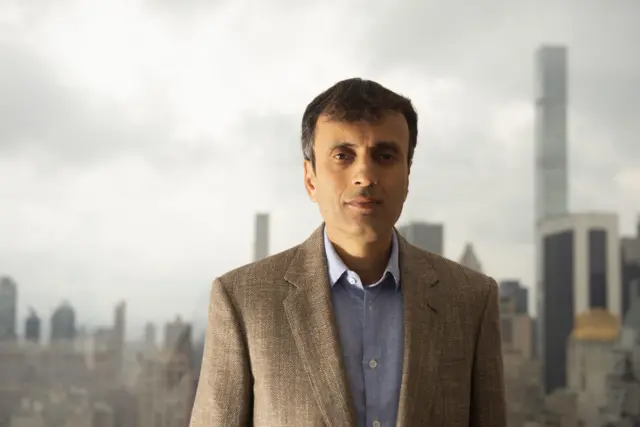 Sharma usando um terno marrom e uma camisa azul clara em baixo. Atrás dele há vários prédios 