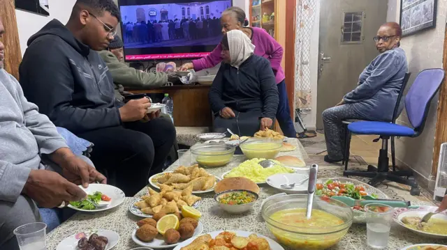 Une table pleine de nourriture se trouve au centre d'un salon et la famille d'Ilham mange autour d'elle.