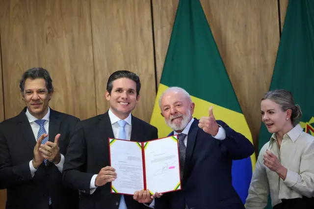 Haddad, Hugo Motta, Lula e Glesi Hoffmann posam para foto em pé; Lula e Hugo Motta seguram documento, enquanto Haddad e Hoffmann aplaudem