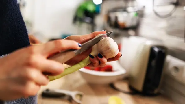 Osoba kroi główkę czosnku trzymaną w ręce, w tle zamazany blat kuchenny
