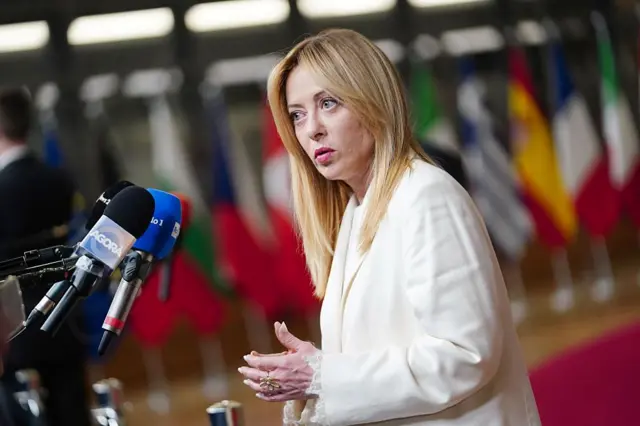 Meloni beyaz bir ceket giymiş. Sol tarafta bulunan mikrofonlara konuşuyor. Arkada pek çok ülkenin yan yana dizilmiş bayrağı flu bir şekilde görülüyor. 