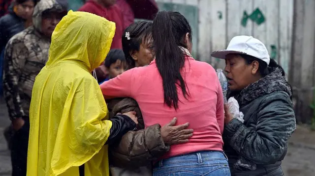 Una mujer, víctima de las inundaciones, es consolada por sus vecinas. Una de ellas tiene un poncho plástico amarillo que le cubre la cabeza en el cuenrpo