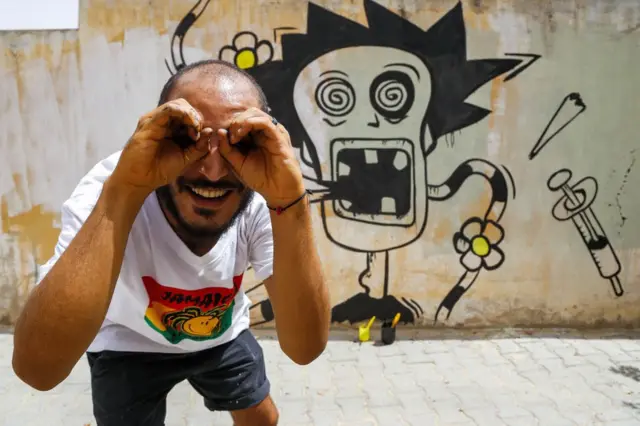 Umuhinga wo muri Tunisia wo gucapa asiga amarangi ku mpome (Graffiti) , Kaisser Grojja, azwi kandi nka Wolf Gang, ariko arifotoza iruhande y'igicapo ciwe ca vuba ku ruhome mu gisagara ca Sidi Bouzid co muri Tunisia ku wa gatanu. 27/08/2021