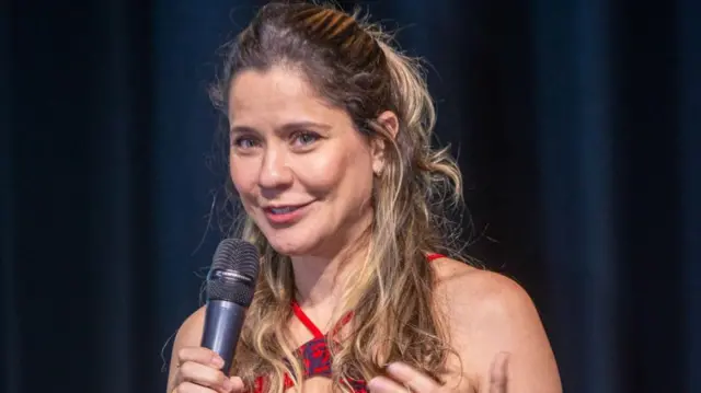 A foto mostra a psicóloga Mayra Gaiato. Ela está com o cabelo preso, tem olhos claros e usa uma blusa roxa. Ela está apoiando o queixo em uma das mãos