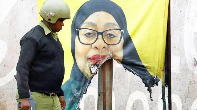 Umupolisi wa Tanzania wo guhosha imvururu ahagaze iruhande rw'ifoto yangijwe ya Perezida Samia Suluhu Hassan yitandiye mu mutwe n'igitambaro cy'umukara, inyuma ye hari ibara ry'umuhondo. Ifoto yo ku itariki ya 30 Ukwakira (10) mu mwaka wa 2025. 