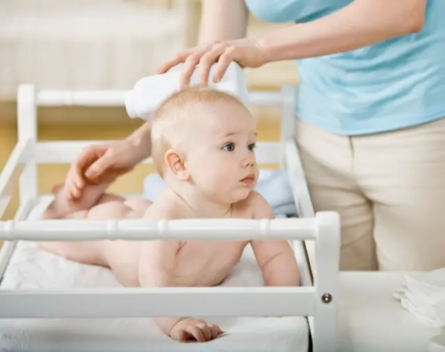 Alt değiştirme masasında yüzüstü yatan bir bebek. Yanındaki yetişkinin elinde bebek pudrası var