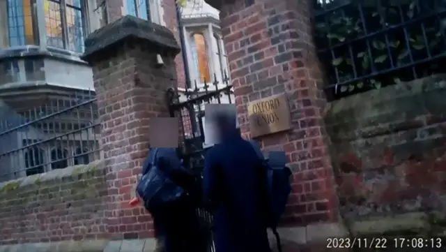 影片畫面顯示兩名面部被模糊處理的男子站在牛津辯論社入口外。他們身穿黑衣，背着背包。