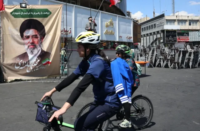 دو دوچرخه سوار در حال عبور از خیابان، تصویری از مجتبی خامنه‌ای نصب شده است و تعدادی صندلی در گوشه‌ای روی هم چیده شده‌اند