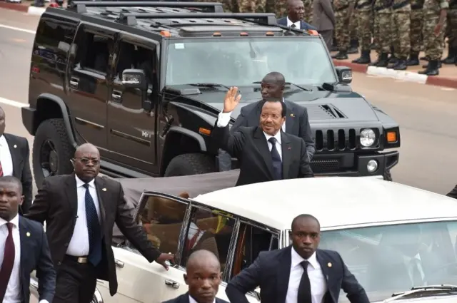 Paul Biya parade dans sa limousine présidentielle, entouré de ses officiers de sécurité. 