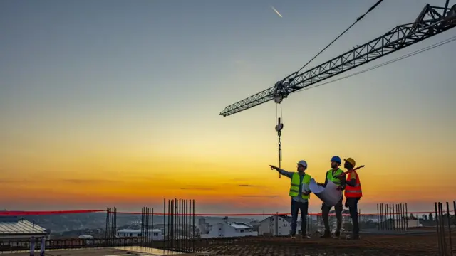 Trois ouvriers du bâtiment en uniforme, debout sur le toit d'un chantier de construction, avec une grue en arrière-plan.