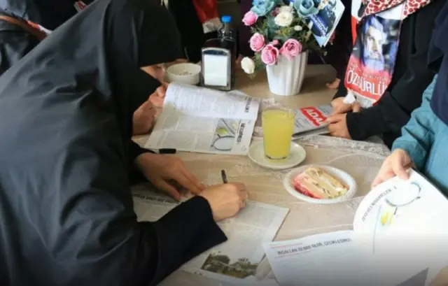 Semra Kuytul bir masada oturmuş, bir derginin üstüne yazı yazıyor. Önünde bir kek ve bir limonata var
