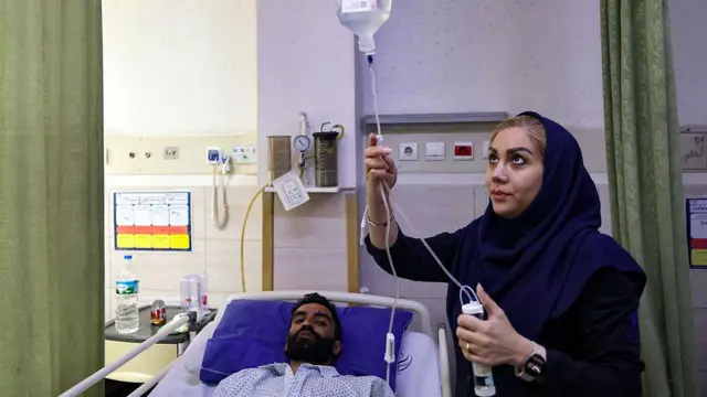 Tahran'daki Rasoul Akram hastanesinde bombalamada yaralanan bir kişi yakını ile bir arada. Adam hasta yatağında yatıyor, başörtülü kadın serumu düzeltiyor.