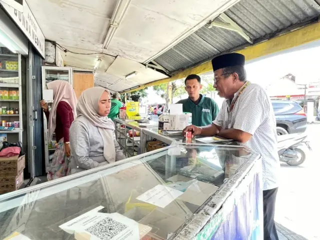 Pengelola apotek di Kota Padang, Debi Septia Nanda sedang nampak berbincang dengan seorang pria tua berkacamata dengan kopiah. Mereka dibatasi dengan etalase setinggi perut dengan latar obat-obatan yang dipajang dalam warung.