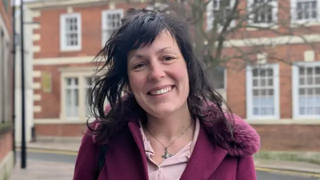 Una persona de cabello oscuro y ondulado se encuentra en una calle tranquila, rodeada de edificios de ladrillo rojo con marcos de ventanas blancos. Lleva un abrigo burdeos intenso con cuello ribeteado de piel sobre una blusa rosa claro y un collar de plata. Detrás de ella se ve un árbol con ramas desnudas.