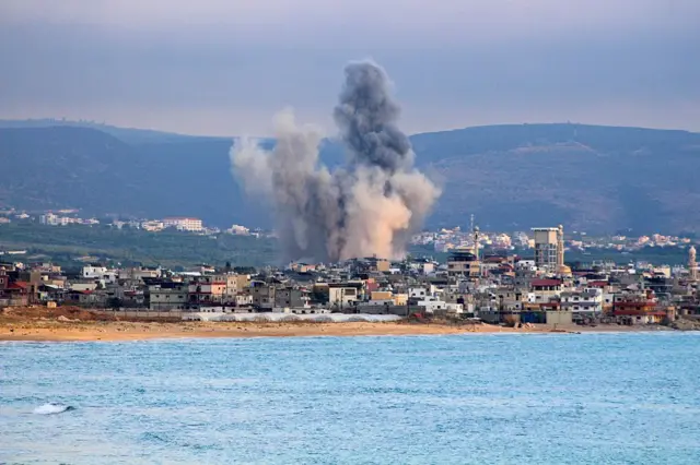 يظهر في الصورة شاطئ عليه عدد كبير من المباني يمتد البحر أمامها بموج هادئ، خلف المباني يتصاعد دخان كثيف عقب غارة إسرائيلية. الصورة التقطت نهاراً.