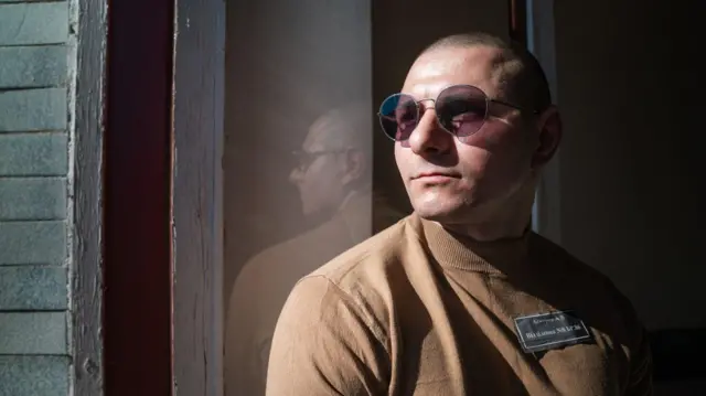 Andrii Askerov, con una camisa marrón y gafas de sol, mira por la ventana de una prisión.
