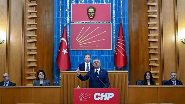 CHP lideri Özgür Özel CHP'nin TBMM grup toplantısında kürsüde konuşuyor. arkasında chp'nin altı oklu amblemi ve Atatürk büstü var. arkada 5 kişi daha oturuyor.