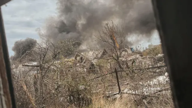Zdjęcie z okna na pustkowie w Pokrowsku. Domy są poważnie uszkodzone, drzewa nagie, a w oddali widać dużą, szaro-czarną chmurę dymu.