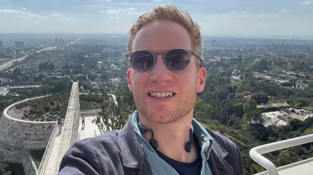 Kieran, con gafas de sol, se toma una selfie desde una terraza elevada al aire libre, con una extensa vista de la ciudad y verdes colinas al fondo. 