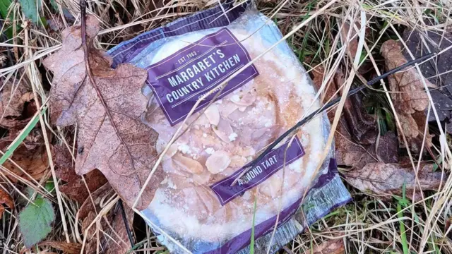 An almond tart, individually wrapped, lying on the grass with dead brown leaves around it and on top of part of the packaging