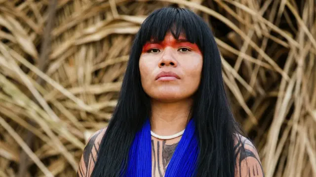 Fotografia colorida mostra uma mulher do povo Yawalapiti, com cabelos lisos e negros, usando uma pintura ritual vermelha e colarcomo sacar o bonus da arbetymiçangas azuis