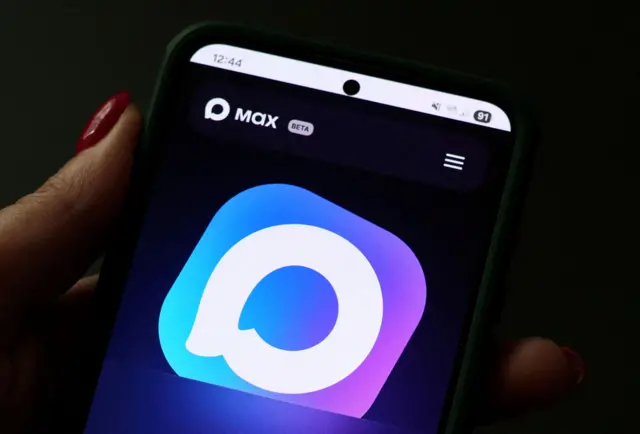 На черном фоне человек держит смартфон левой рукой. Виден большой палец с красным лаком. На смартфоне видно время 12:44 и изображение светлофиолетового квадрата с закругленными углами В квадрате находится кружок изображающий сообжение. В левом верхнем углу экрана написано Max Beta.