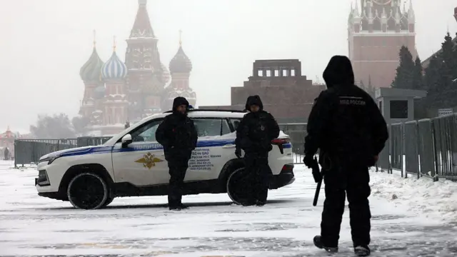 Заснеженная Красная площадь в Москве. На переднем плане – трое вооруженных полицейских рядом с белым внедорожником. В глубине кадра видны собор Василия Блаженного, мавзолей Ленина и башня Кремля с часами. Погода пасмурная, моросящий снег, ограждения ограничивают движение прохожих.