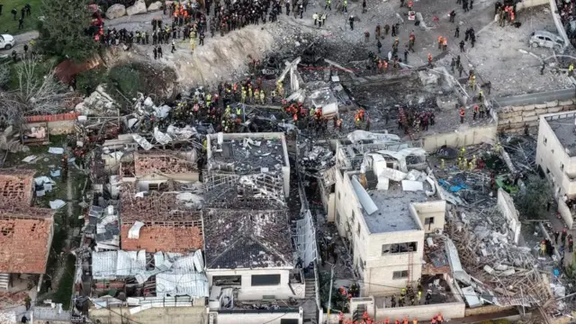 立ち並ぶ複数の建物が破壊され、周りに多くの作業員が立っている様子のドローン写真