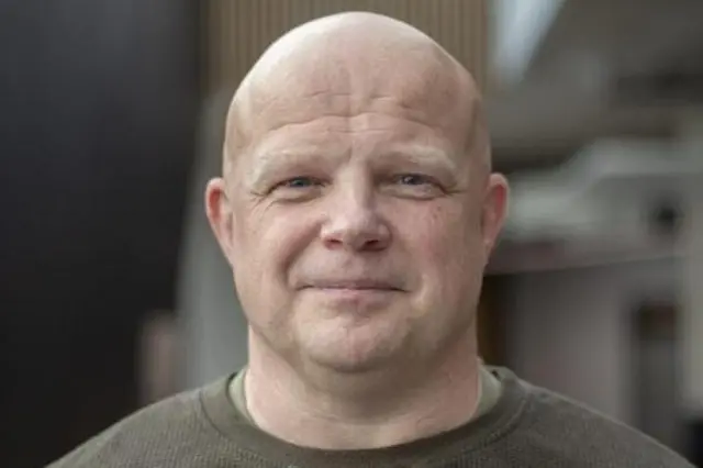 Endru Bartlet u centru fotografije se osmehuje u kameru. On je srednjih godina, ćelav i ima plave oči. Nosi kaki majicu dugih rukava.