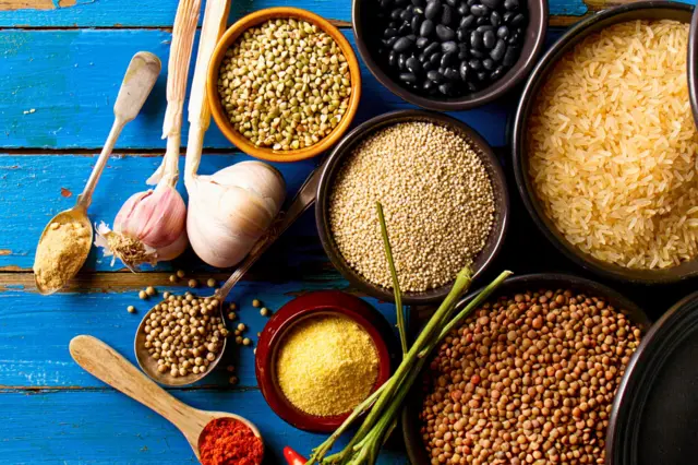 pişmemiş pirinç, mercimek, siyah fasukye, kinoa, karabiber, kırmızı biber hepsi ayrı kaplarda ve mavi bir tahta masa üzerinde duruyor.