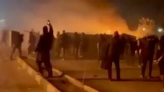 ویدیویی تار که معترضان و دود ناشی از آتش‌سوزی را نشان می‌دهد و فردی در حال فیلم‌برداری با تلفن همراه است؛ مشهد، اوایل ژانویه