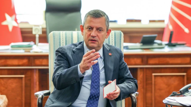 CHP lideri Özgür Özel makamında elinde notlarla fotoğrafta olmayan birilerine konuşuyor Elini havaya kaldırmış durumda, arkada CHP ve Türk bayrakları var.