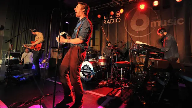 La banda Kaiser Chiefs tocando en un pequeño escenario interior, con una cortina con el logo de BBC Radio 6 Music detrás de ellos.