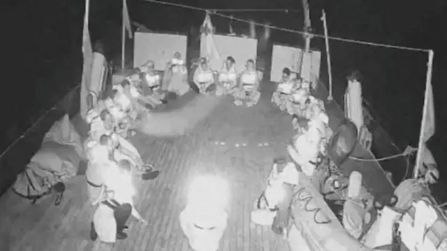 Siyah beyaz bir gece görüşü kamerasından geminin güvertesinde daire şeklinde oturmuş aktivistler görünüyor. 
