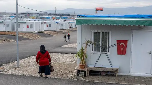 Üzerinde yer yer Türk bayrakları olan konteyner'ların olduğu alanda bir kadn yürüyor, ileride sokak ortasında iki çocuk seçiliyor. 