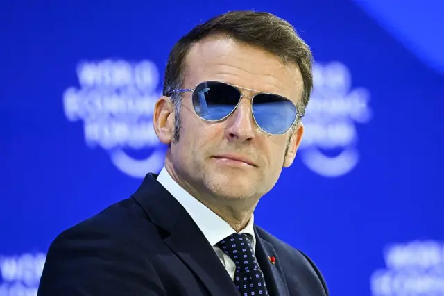 Macron mavi parlayan bir güneş gözlüğü takıyor. Takım elbisesi var ve arkada mavi fonda konuşuyor